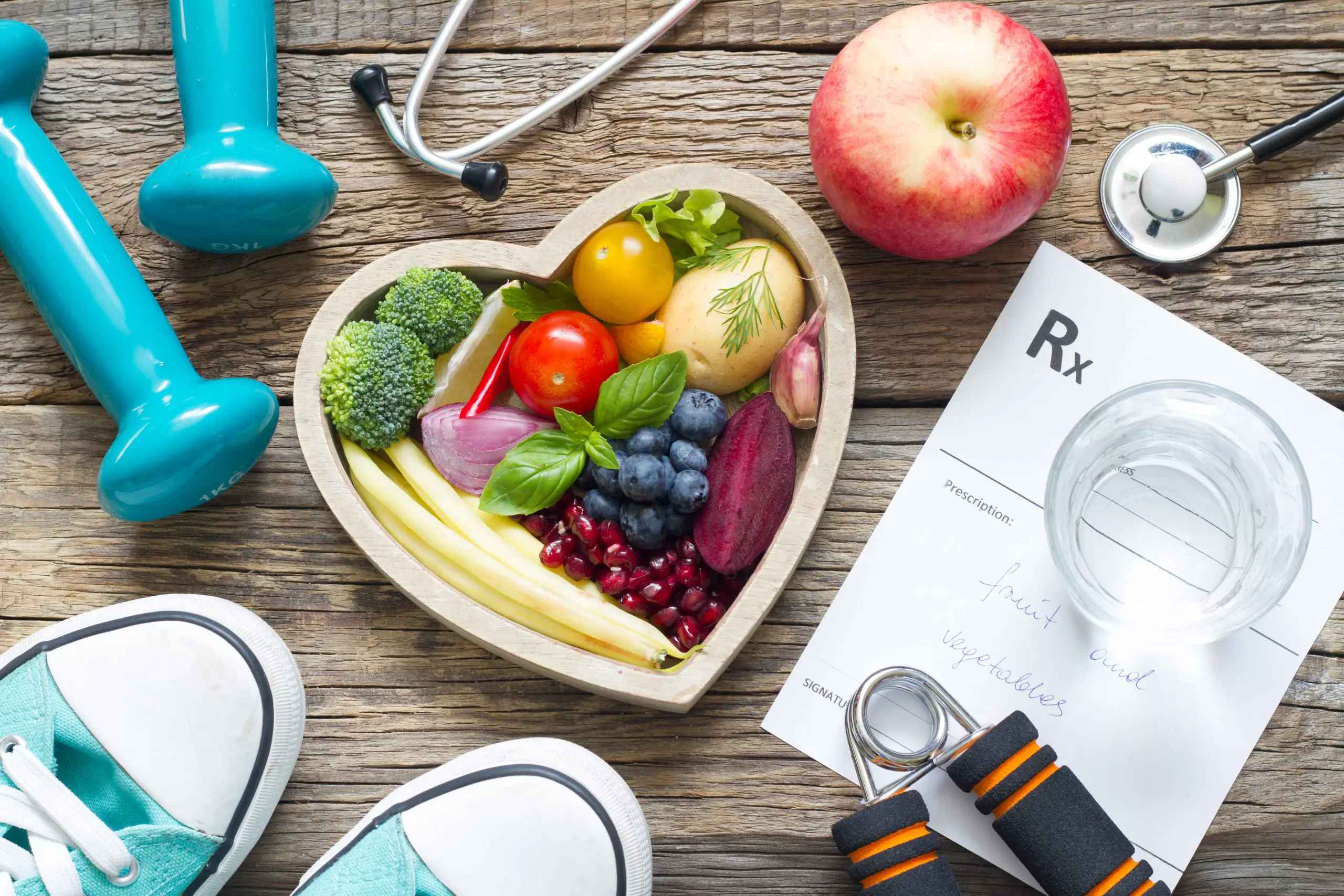 Harmony Hormone Solutions - Heart-shaped bowl of fruits and vegetables, dumbbells, stethoscope, red apple, sneakers, a prescription note, hand gripper, and glass of water on a wooden surface.