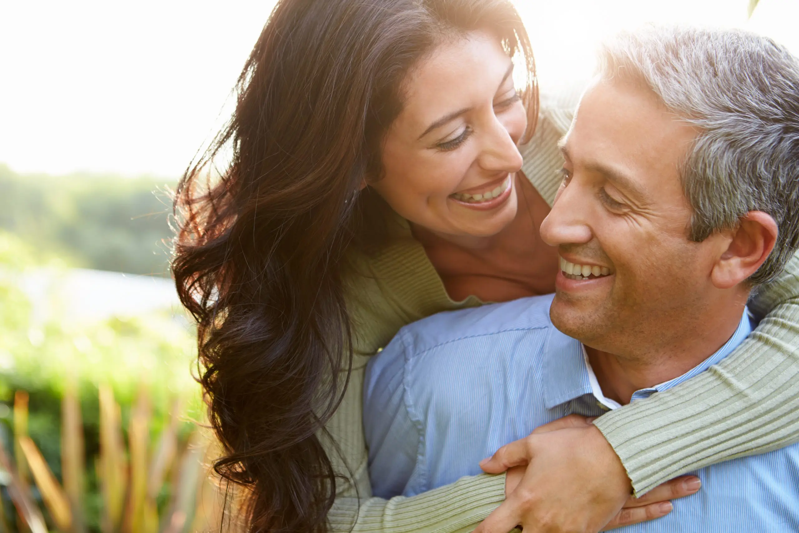 Harmony Hormone Solutions - A woman with long hair embraces a smiling man from behind outdoors in a sunny, green setting.