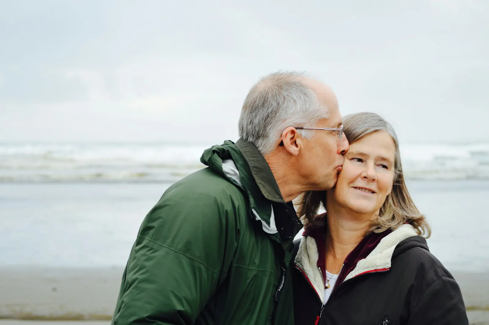 Harmony Hormone Solutions - An older man kisses a woman on the cheek as they stand on a breezy beach, both wearing jackets. Their comfortable closeness feels like a testament to enduring love, much like navigating menopause symptoms with understanding and care.