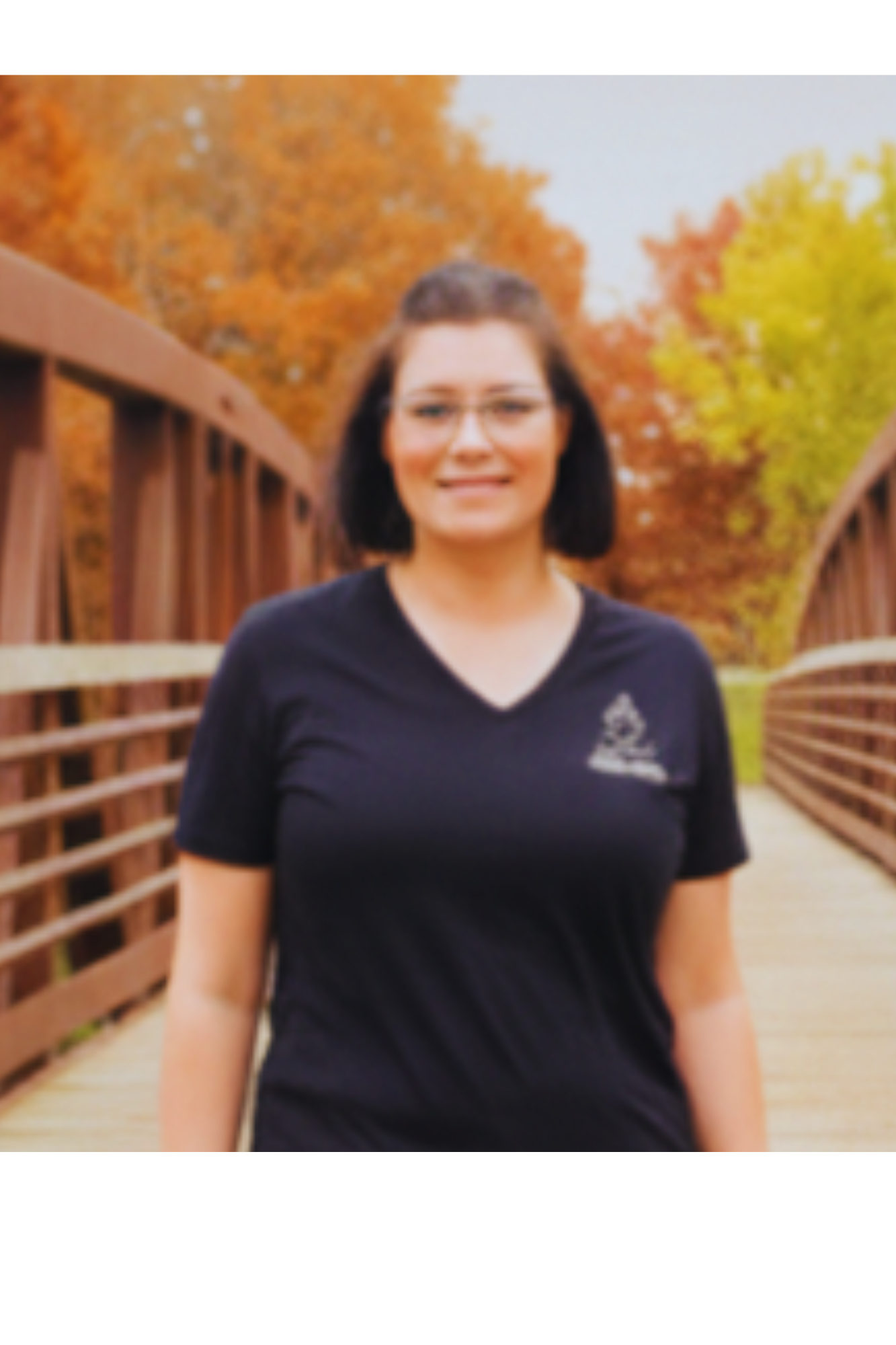 Harmony Hormone Solutions - A woman with short dark hair and glasses stands on a wooden bridge with autumn trees in the background.