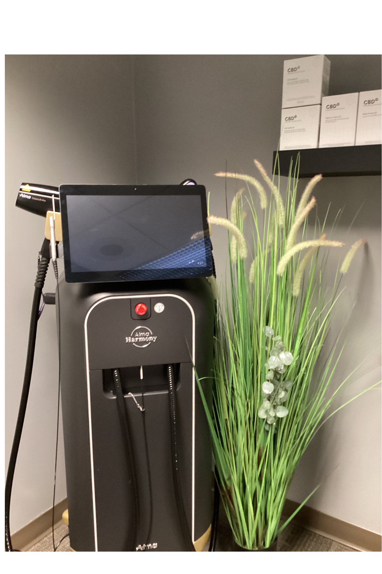 Harmony Hormone Solutions - A black medical aesthetic device with a touch screen stands next to a decorative green grass plant and a shelf with white product boxes against a gray wall.