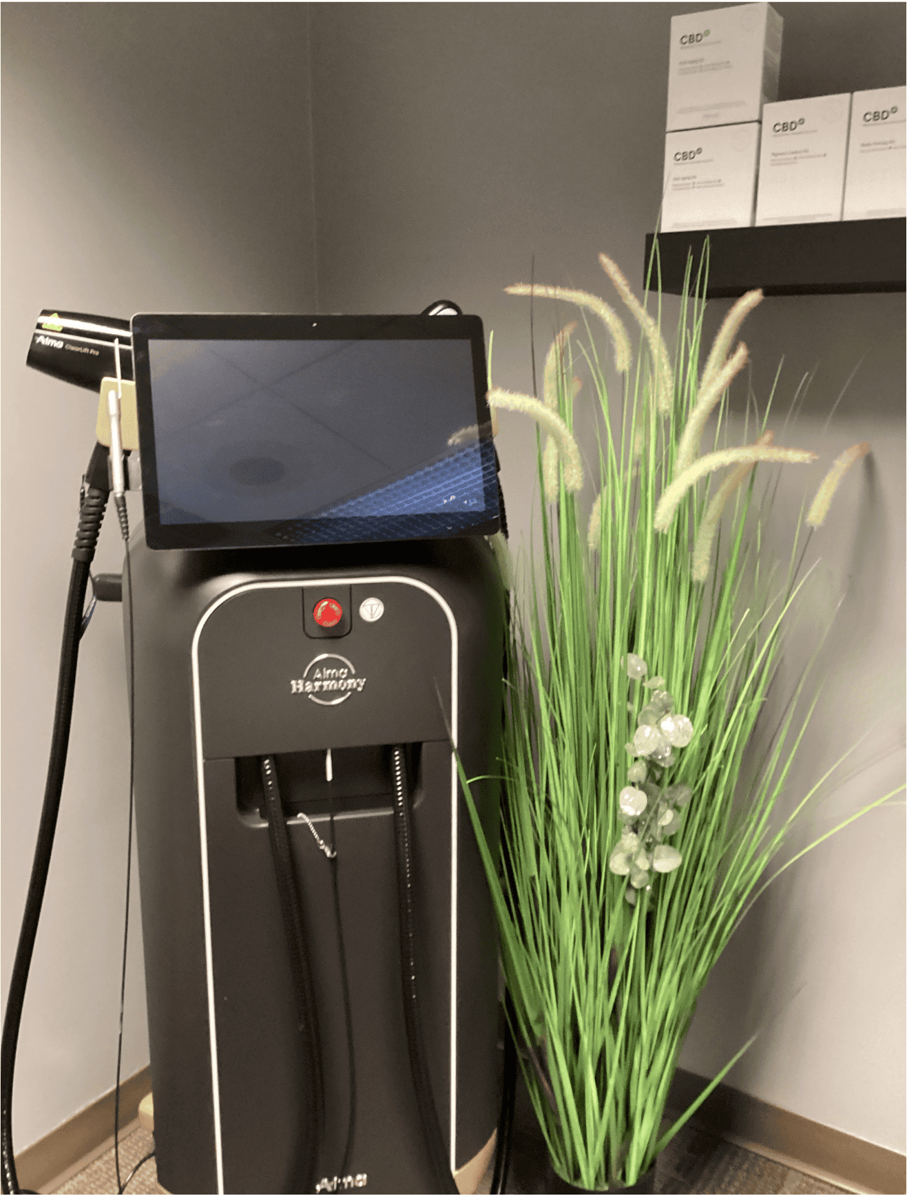 Harmony Hormone Solutions - A black medical aesthetic device with a touch screen stands next to a decorative green grass plant and a shelf with white product boxes against a gray wall.